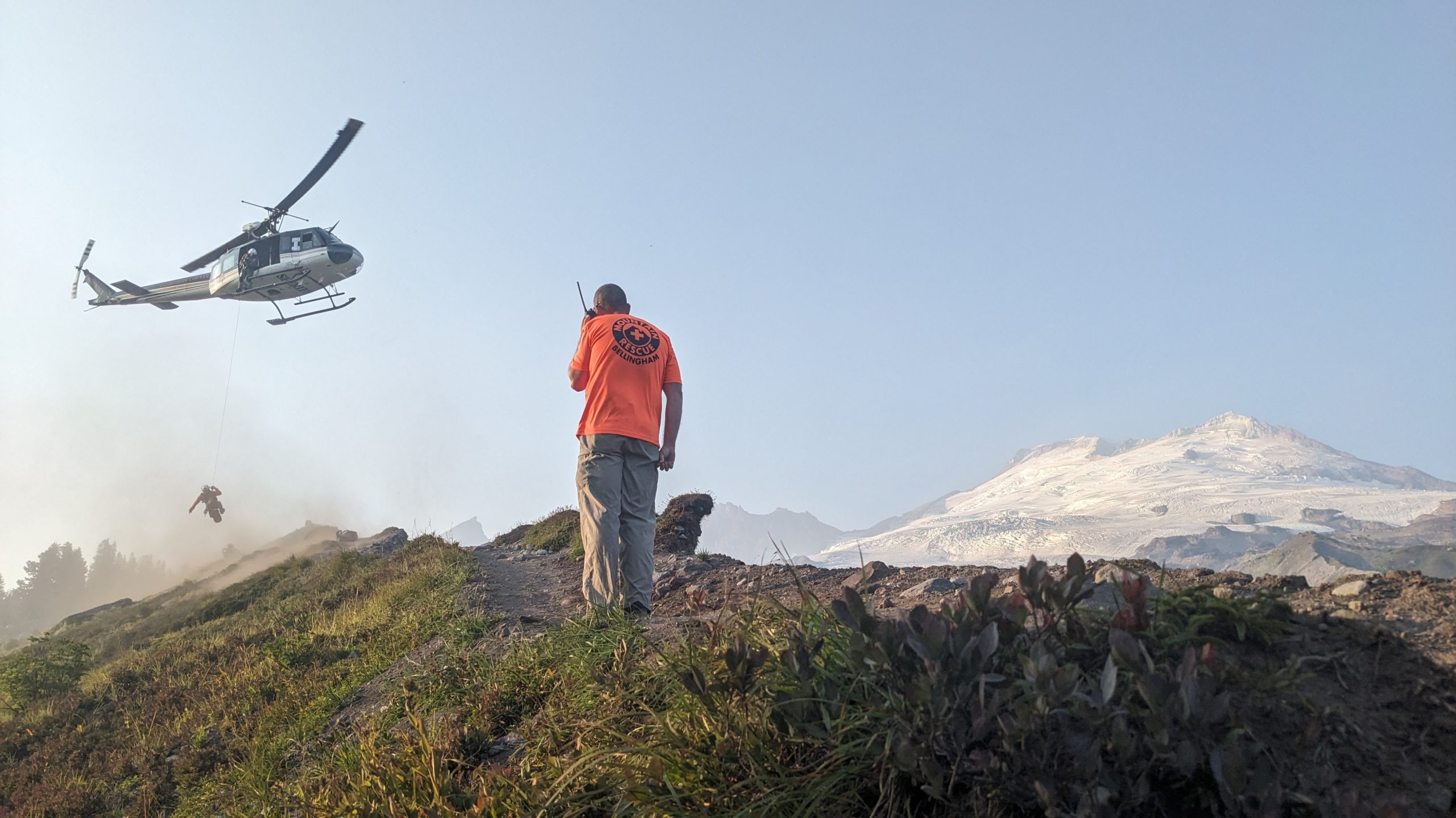 Helicopter flying in late summer with a rescuer being hoisted inside with Mount Baker in the background and another rescuer communicating in the foreground via radio.